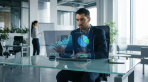 Man analyzing financial dashboards on a transparent screen in a modern, sunlit office. Graphs show growth; a colleague is in the background.
