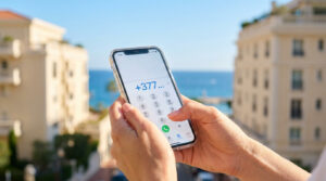 Mains composant l'indicatif +377 sur un smartphone, avec vue sur les bâtiments de Monaco et la mer Méditerranée.